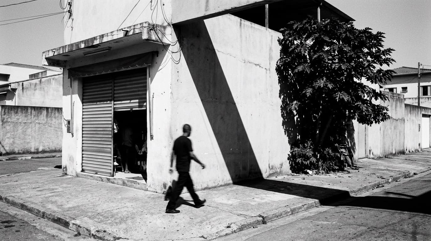 Esquina de um bairro com uma loja de porta de enrolar semiaberta e uma pessoa atravessando em movimento